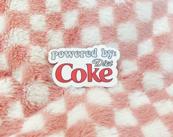 Può includere: Un adesivo bianco con la scritta "powered by: Diet Coke" in un font retrò. La parola "Coke" è rossa, bordata di bianco. L'adesivo è su uno sfondo a scacchi rosa e bianco.