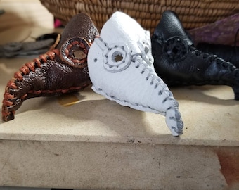 May include: Three leather plague doctor masks, one brown, one white, and one black. The masks are stitched with a contrasting thread and have a long beak.