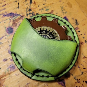 May include: A round, green leather pouch with a brown gear-shaped object inside. The pouch has black stitching around the edges. The gear has numbers and letters on its face. The leather has a distressed appearance.