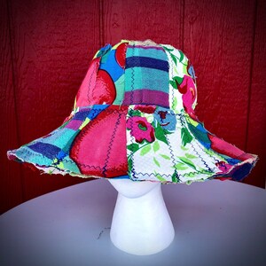 May include: A bucket hat constructed from various fabric patches featuring vibrant colors including bright pink, blue, green, and white. The hat displays a mix of floral and plaid patterns joined by visible dark zigzag stitching along the seams.