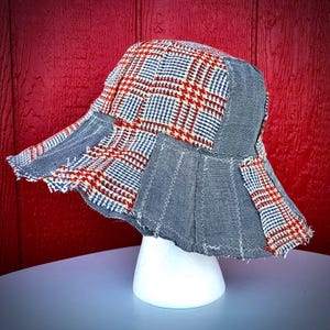 May include: A bucket hat constructed from a patchwork of red, white, and blue plaid fabric panels interspersed with solid grey denim panels. The hat features visible white zigzag stitching along the seams and a frayed, raw edge along the brim.