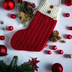 May include: Red and white crocheted Christmas stocking with a white cuff and two brown buttons. The stocking is surrounded by red and gold ornaments and a small evergreen sprig.