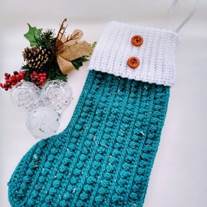 May include: A teal crocheted Christmas stocking with a white crocheted cuff and two brown buttons. The stocking has a white loop at the top.