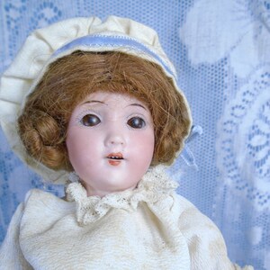 Könnte beinhalten: Eine Vintage-Porzellanpuppe mit braunen Haaren und Augen, gekleidet in einer cremefarbenen Bluse mit Spitzenbesatz, einem schwarzen Samtrock und einer Haube mit blauem Band. Das Gesicht der Puppe ist bemalt mit rosigen Wangen und einem kleinen, offenen Mund.