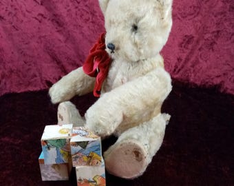 Puede incluir: Un oso de peluche blanco vintage con un lazo rojo se sienta sobre una superficie burdeos y juega con un conjunto de bloques de madera coloridos con ilustraciones.