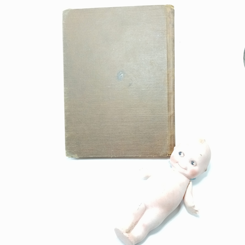 May include: A vintage book with a textured, tan cover is positioned next to a small, porcelain Kewpie doll. The doll has a smiling expression and is lying on its back. The book's spine is visible, and the pages are bound together.