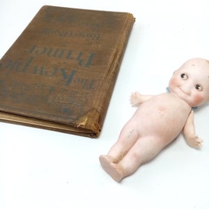 May include: A vintage brown book titled "The Kewpies Primer" by Rose O'Neill, next to a porcelain Kewpie doll with a blue ribbon. The book has a worn cover. The doll has rosy cheeks and a smiling expression.