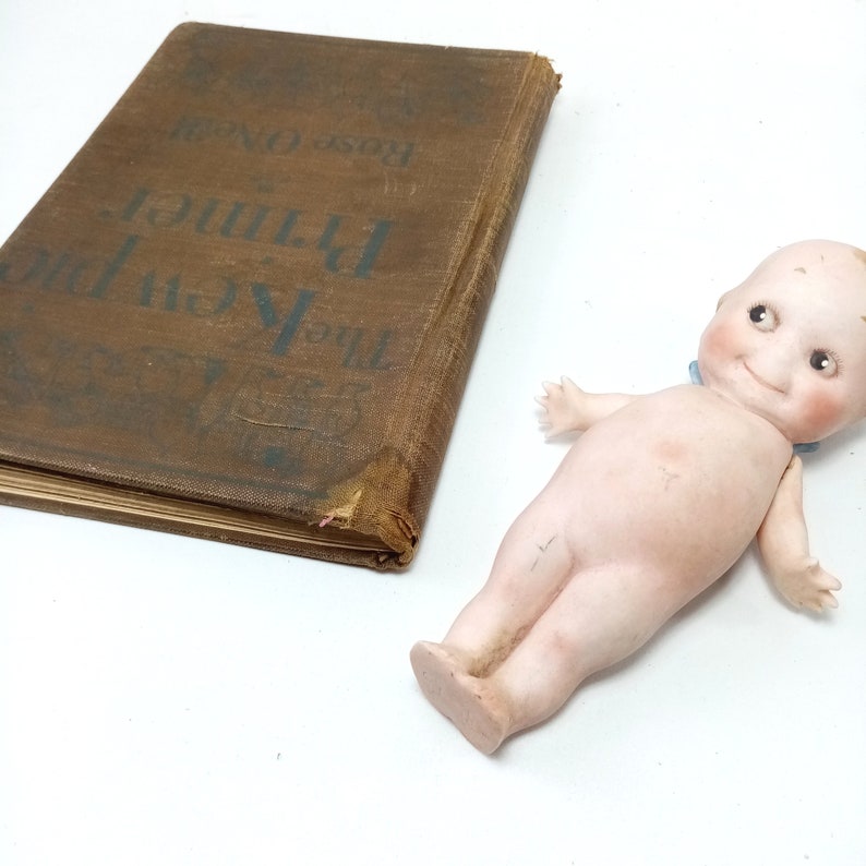 May include: A vintage brown book titled "The Kewpies Primer" by Rose O'Neill, next to a porcelain Kewpie doll with a blue ribbon. The book has a worn cover. The doll has rosy cheeks and a smiling expression.