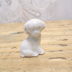 May include: A small, white ceramic dog figurine with a rounded head and a seated posture. The figurine has subtle brown speckles and is set against a light-colored wooden surface.