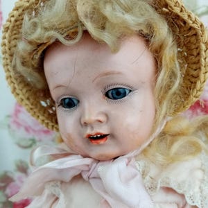 May include: A vintage doll with blue eyes, blonde curly hair, and a straw hat. The doll is wearing a pink dress with a matching bow. The doll's face has a painted expression with red lips.
