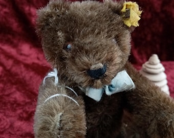 Pode incluir: Um urso de peluche castanho com uma gravata borboleta azul e um botão amarelo na orelha. O urso está sentado numa superfície de madeira.