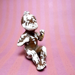 May include: A small, vintage doll figurine in a seated position. The doll is painted brown with white paint chipping off, revealing the base material. The background is a pink and white striped pattern.
