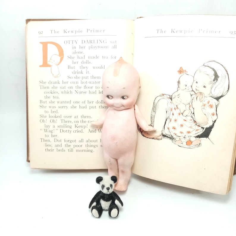 May include: A vintage Kewpie doll stands next to an open book titled "The Kewpie Primer." The doll is nude with a painted face and a small panda bear figurine is in front of the book. The book pages display illustrations and text.