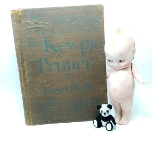 May include: An antique book titled "The Kewpie Primer" by Rose O'Neill, with a brown cover and teal lettering, stands next to a vintage Kewpie doll and a small panda bear figurine. The doll is pale with a distinctive hairstyle.