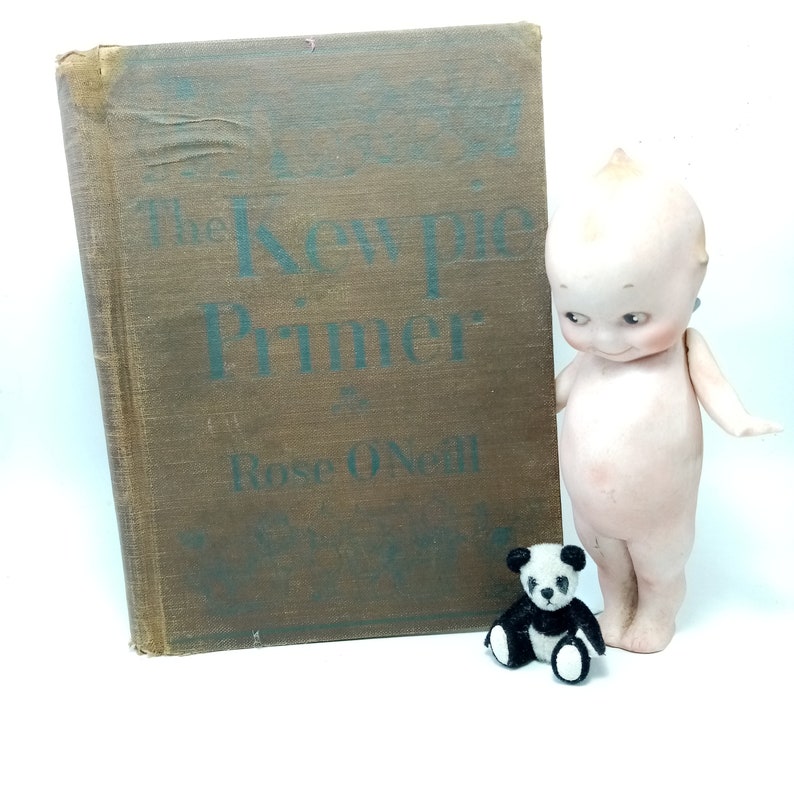 May include: An antique book titled "The Kewpie Primer" by Rose O'Neill, with a brown cover and teal lettering, stands next to a vintage Kewpie doll and a small panda bear figurine. The doll is pale with a distinctive hairstyle.