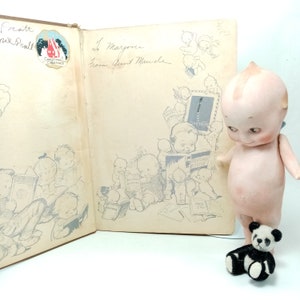 May include: An open vintage book with illustrations of babies reading, a Christmas greeting, and handwritten text. A porcelain doll and a small panda bear toy are next to the book.