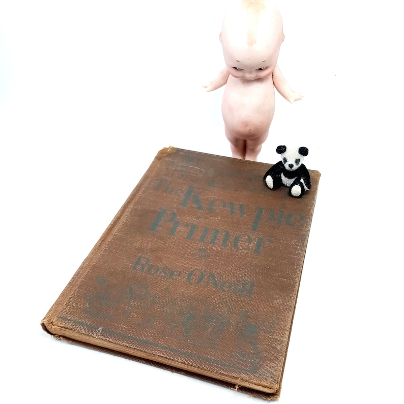 May include: A vintage brown book titled "The Kewpie Primer" by Rose O'Neill, with a small panda figurine and a Kewpie doll. The book has faded green text and is set against a white background.