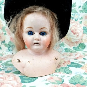 May include: Antique doll head with blue eyes, blonde hair, and a black hat. The doll's face has rosy cheeks and a serene expression. The porcelain head is a vintage collectible.
