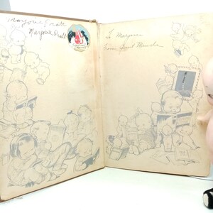 May include: An open vintage book with illustrations of babies reading. The left page has the names "Marjorie Pratt" and a Christmas Greetings sticker. The right page has a handwritten message. The book is brown with a light tan interior.