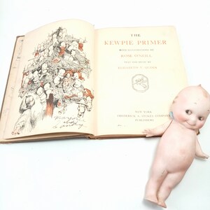 May include: An open vintage book titled "The Kewpie Primer" with illustrations by Rose O'Neill. The book features a drawing of a tree made of Kewpie dolls. A porcelain Kewpie doll with blue wings rests beside the book.