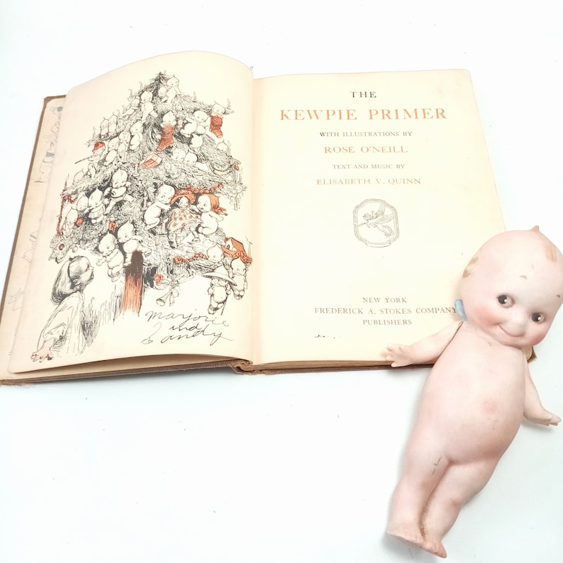May include: An open vintage book titled "The Kewpie Primer" with illustrations by Rose O'Neill. The book features a drawing of a tree made of Kewpie dolls. A porcelain Kewpie doll with blue wings rests beside the book.