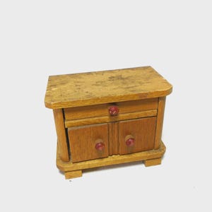 May include: A miniature wooden cabinet featuring a rectangular top surface with a weathered finish, one horizontal drawer, and two lower cabinet doors. Each drawer and door is fitted with a small, round, dark red knob for opening and closing.