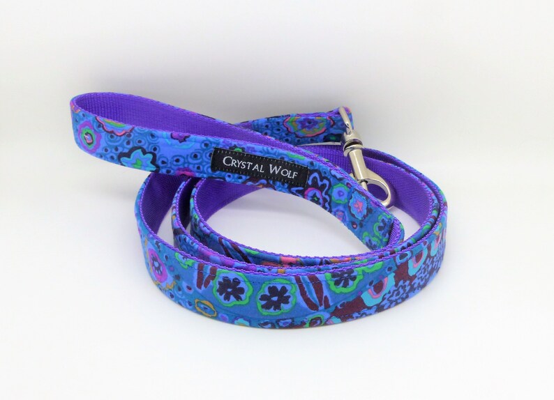 May include: A blue dog leash with a vibrant floral pattern in shades of blue, purple, green, and pink. The leash has a purple handle and backing, with a silver metal clasp. The brand name "CRYSTAL WOLF" is printed on a black label.