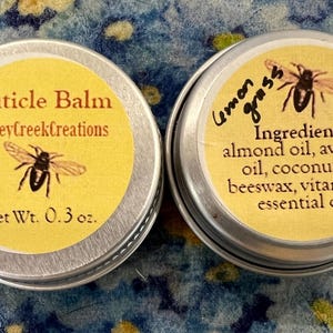 Op de afbeelding: Twee kleine, zilveren blikjes met gele etiketten. Het blikje aan de linkerkant is gelabeld "Cuticle Balm HoneyCreekCreations Net Wt. 0.3 oz." en heeft een afbeelding van een zwarte en gele bij. Het blikje aan de rechterkant is gelabeld "Lemon Grass" en vermeldt de ingrediënten: "amandel olie, avocado olie, kokosolie, bijenwas, vitamine E, etherische olie."