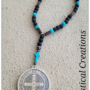May include: A silver St. Benedict medal with a turquoise cross and black beads on a necklace. The medal has the inscription "S.B. Pax. V.B. N.D.S.M.D. P.M.L.B. A.M.S.N. Italy".