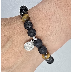 May include: A beaded bracelet featuring alternating black porous lava stone beads, smooth dark gray hematite beads, and golden-brown tiger eye beads. A circular silver-toned metal charm with an embossed cross design hangs from the bracelet.