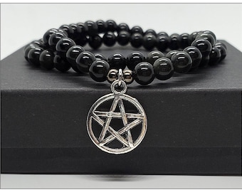 Puede incluir: Pulsera de cuentas negras con un dije de pentagrama plateado. La pulsera presenta dos hilos de cuentas negras redondas y pulidas. El dije plateado es una estrella de cinco puntas dentro de un círculo.