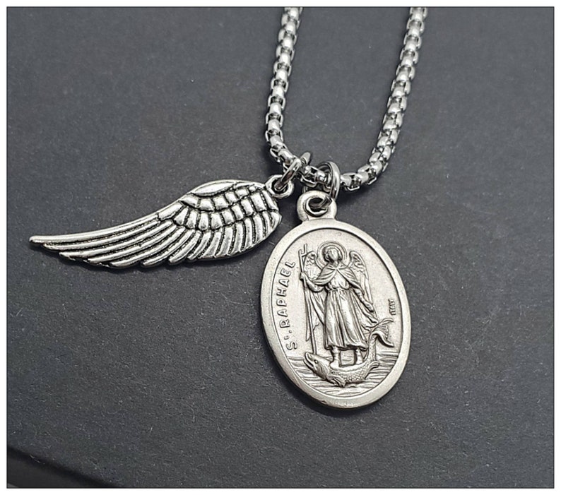 May include: Silver chain necklace with a pendant featuring a depiction of Saint Raphael and an angel wing charm. The pendant is oval shaped and has the words "S. RAPHAEL" inscribed on it.