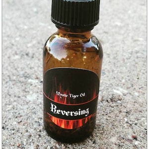 May include: A small amber glass bottle with a black cap. The label reads "Mystic Tiger Oil" above the word "Reversing" in a stylized font, with a fiery red and orange graphic.