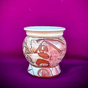 May include: A handcrafted ceramic pot with a wide, rounded body and a pedestal base. The pot features a cream-colored rim and base, with a design of reddish-brown abstract shapes on a white background. The background is a vibrant magenta.