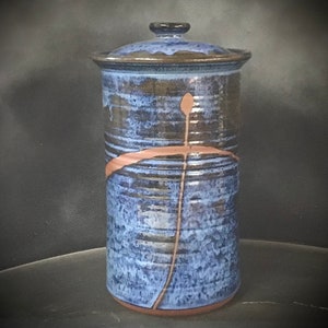 May include: A tall cylindrical ceramic canister with a lid, featuring a mottled blue and dark brown glaze. A thin, unglazed terracotta-colored line forms a cross-like pattern across the front surface, contrasting with the textured blue finish.