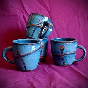 May include: Four blue ceramic mugs with brown line designs. The mugs have a curved handle and a glossy finish. The mugs are stacked on a purple background. Each mug is approximately 10 cm in height.