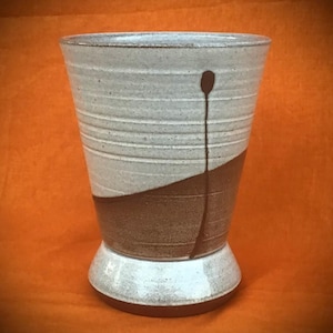 Könnte beinhalten: Eine handgefertigte Keramikvase mit einem einzigartigen Design. Der obere Teil ist hellgrau mit horizontalen Linien, während die untere Hälfte braun ist. Ein dunkelbrauner, tropfenförmiger Akzent ist auf die Seite gemalt. Die Vase steht auf einem weißen Sockel.