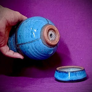 Può includere: Un contenitore in ceramica blu, rotondo, con una smaltatura testurizzata e maculata e una base marrone scuro non smaltata che presenta una piccola apertura circolare e una firma scritta a mano. Un coperchio in ceramica blu abbinato è posato accanto.
