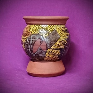 May include: A round terracotta vessel featuring a wide rim and a flared base. The exterior surface is decorated with a pattern of small yellow dots and dark gray abstract shapes, contrasting with the natural reddish-brown clay of the rim and base.