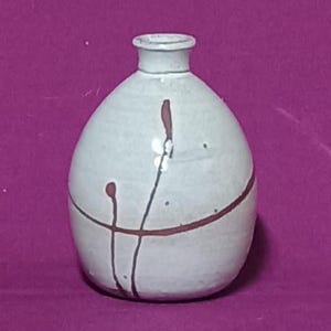 Può includere: Piccolo vaso in ceramica bianco sporco con corpo arrotondato e collo stretto. Decorato con linee e punti rosso scuro, creando un design astratto. Il vaso ha una finitura liscia e opaca ed è probabilmente fatto a mano.