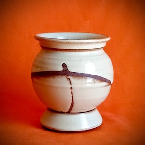 May include: A handcrafted, off-white ceramic vase with a rounded body and a pedestal base. The vase features a brown horizontal band and a vertical brown line, adding a touch of rustic charm. The vase is suitable for home decor.