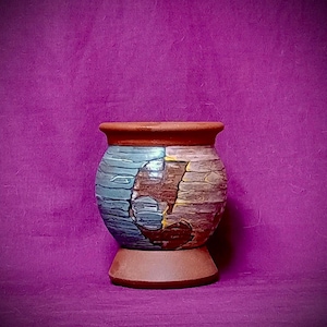May include: A handcrafted ceramic vase with a textured surface. The vase features a color palette of blue, brown, and tan, with a reddish-brown rim and base. The design is abstract and artistic, suitable for home decor.