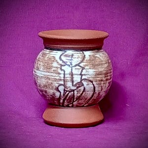 Peut inclure: Un petit récipient en céramique de couleur terre cuite, rond, avec un bord et une base évasés. Le corps présente une finition texturée à rayures horizontales dans des tons marron et blanc, avec un dessin abstrait sombre tracé à la main.