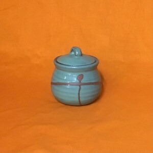 May include: A small, round, ceramic jar with a lid. The jar is a muted teal color with a brown band and a vertical line design. The lid has a small handle. The jar is set against an orange background.