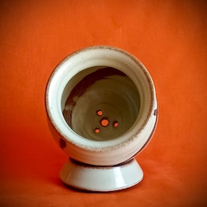May include: A ceramic salt cellar with a white exterior and a brown interior. The salt cellar has a round shape with a pedestal base and three small holes at the bottom. The background is a solid orange color.