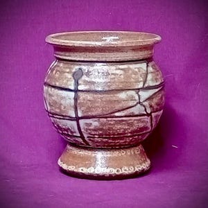 Peut inclure: Un récipient en céramique avec un corps arrondi et un bord évasé, présentant un vernis marron et beige tacheté. La surface est décorée de fines lignes sombres entrecroisées formant un motif géométrique, avec une bordure pointillée texturée.