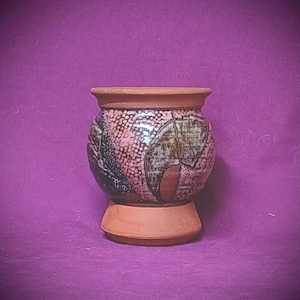 Puede incluir: Un recipiente de cerámica redondo con un borde y una base de terracota sin esmaltar. El cuerpo central presenta un esmalte brillante multicolor con un patrón texturizado de pequeños puntos y formas abstractas oscuras en tonos rosa, marrón y negro.