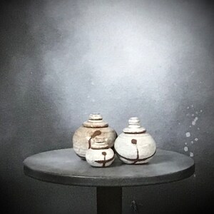 May include: Three white ceramic jars with brown accents, arranged on a gray table. The jars have a rounded shape and a lid. The jars are decorated with simple brown lines and dots.