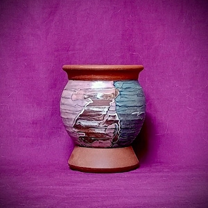 May include: A handcrafted ceramic vase with a unique design. The vase features a round body with a textured surface, showcasing a blend of colours including brown, blue, and lavender. It has a wide, reddish-brown rim and sits on a matching base. The vase is approximately 15 cm tall.