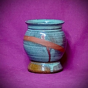 May include: A ceramic cup with a rounded body and a flared rim. The exterior features a mottled blue-grey glaze with a prominent diagonal terracotta-colored stripe and a solid brown base. The vessel has a wide opening and a sturdy, circular foot.